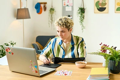 Jeune graphiste blanche concentrée en train de dessiner de la main droite sur une tablette et regardant son ordinateur. Son avant-bras gauche, posé sur le bureau est remplacé par une prothèse.